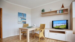 Una sala de estar con piso de madera, una mesa de comedor con sillas, un televisor sobre un mueble, un cuadro de un barco y una estantería con libros y una planta.