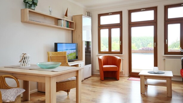 Una sala de estar moderna con piso de madera, televisor, mesa de comedor con sillas y ventanas amplias que ofrecen vistas a la vegetación.