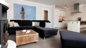 Un salon moderne avec un canapé d’angle noir, une table basse en bois et un fauteuil noir. On aperçoit une cuisine à l’arrière-plan, avec des meubles blancs et une hotte de cuisinière en acier inoxydable.