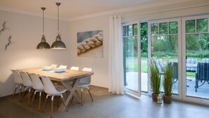 Un coin repas avec une table en bois et des chaises blanches, une grande fenêtre donnant sur un jardin et deux lampes suspendues.