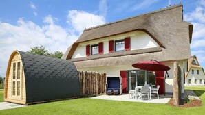 Une maison avec un grand toit de chaume, une remise en bois et une terrasse aménagée avec du mobilier d’extérieur.