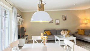 Un coin repas avec une table blanche, des chaises et un vase rempli de fleurs jaunes. Une lampe blanche est suspendue au-dessus de la table, et un canapé avec des coussins jaunes se trouve à l’arrière-plan.