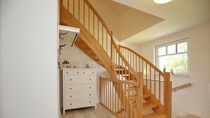 Un escalier en bois avec une rampe, une commode blanche à tiroirs et une fenêtre donnant sur un paysage verdoyant.