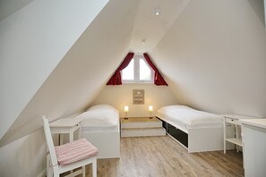 Um quarto sótão pequeno e triangular com duas camas de solteiro, uma escrivaninha e uma cadeira.