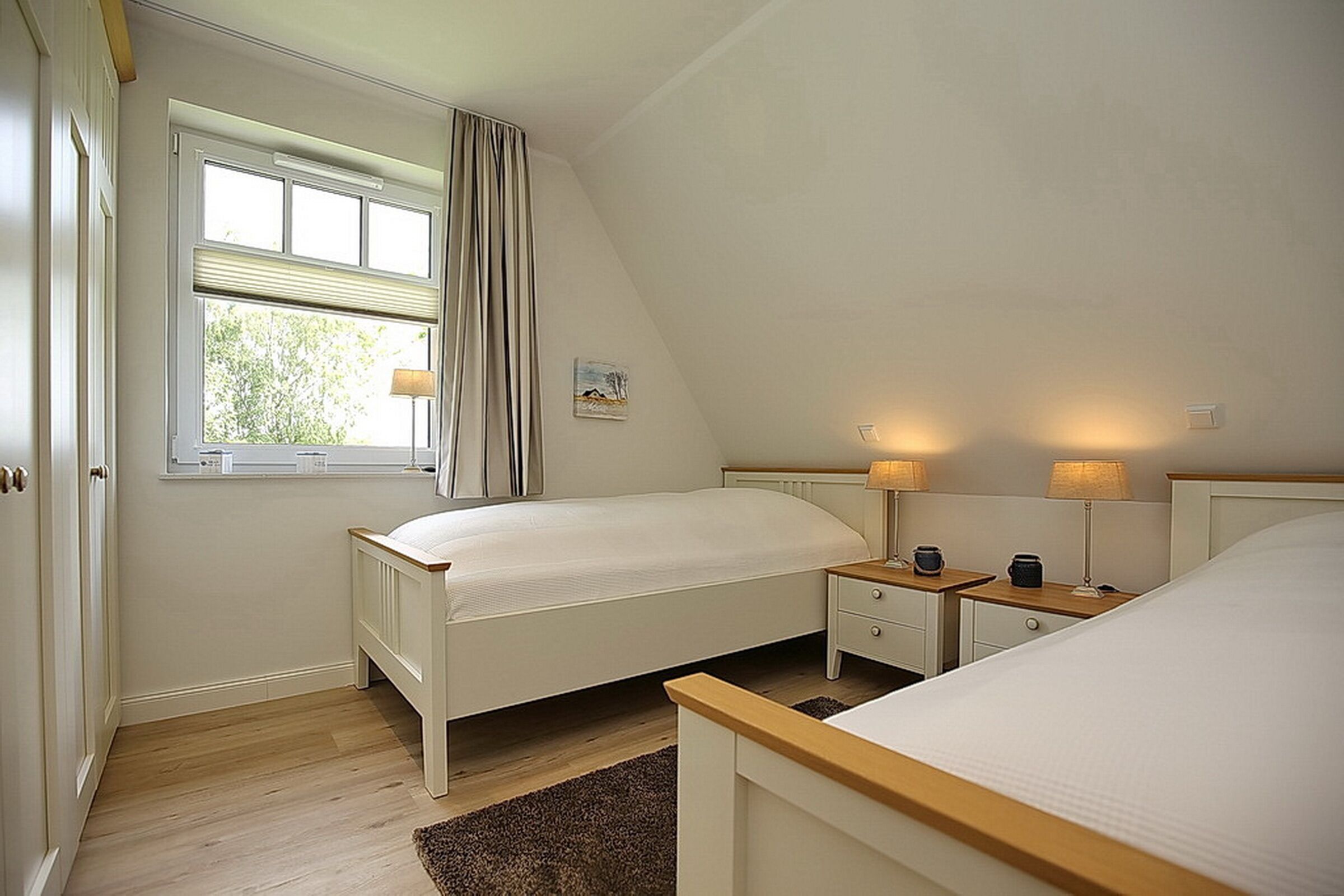 A bedroom with two beds, a window with curtains, bedside lamps, and a small painting on the wall.