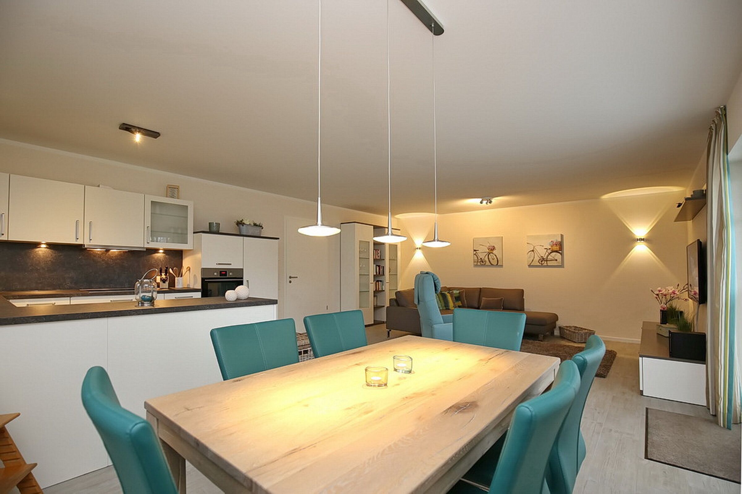 A modern kitchen with white cabinets, a wooden dining table, teal chairs, pendant lights, and a living area with a sofa and a TV.