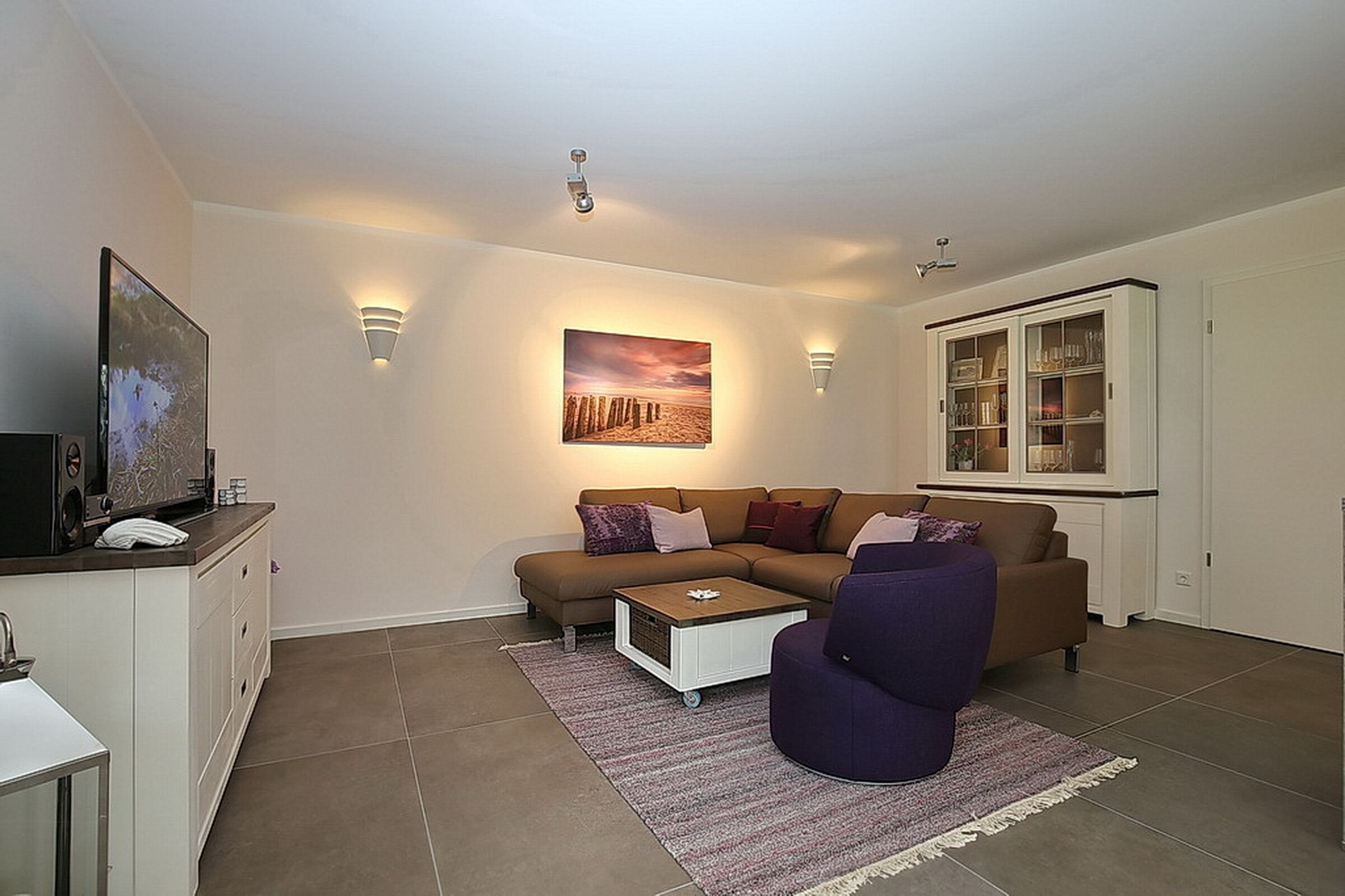 A living room with a brown sofa, a purple armchair, a coffee table, a cabinet with glass doors, and a painting on the wall.