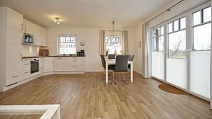 Una cocina moderna con armarios blancos, un comedor con mesa y sillas, y ventanas grandes con persianas.
