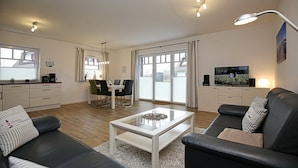 Una sala moderna con sofá, mesa de centro y televisor. Hay un comedor con mesa y sillas, y un área de cocina con armarios y electrodomésticos.