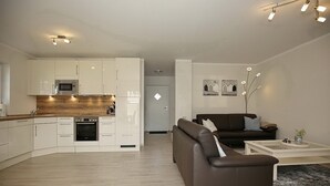 Cozinha moderna com armários brancos, micro-ondas e forno. Na sala de estar, há um sofá de couro marrom e uma mesa de centro.