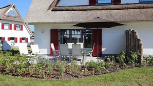 Uma casa com telhado de palha, paredes brancas e persianas vermelhas. Possui um alpendre com mobiliário de exterior e um grande guarda-sol.