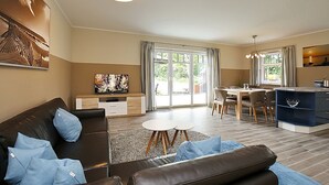 Uma sala de estar moderna com sofá, mesa de centro e televisão. Há também uma área de refeições com cadeiras e uma mesa.