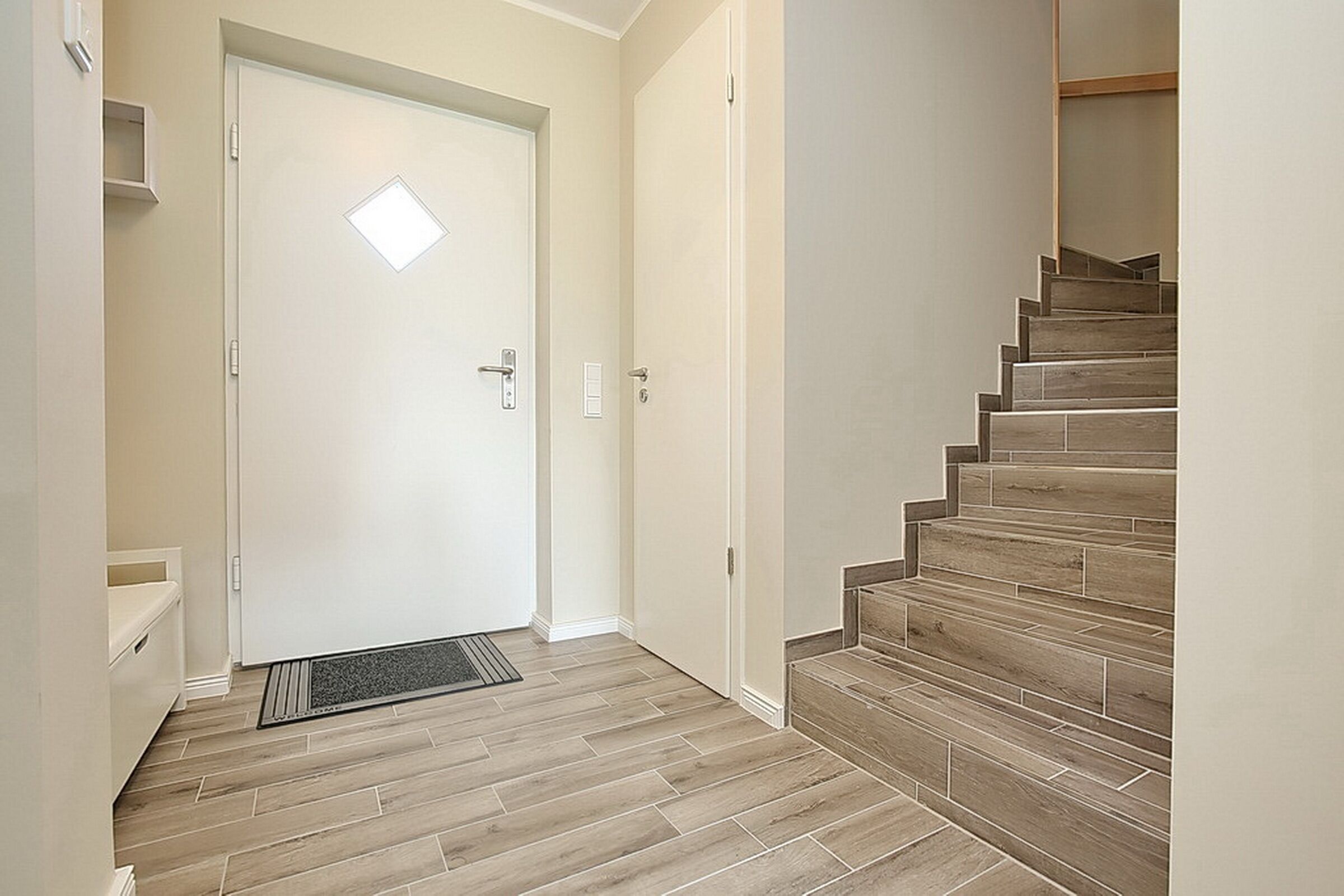 Un pasillo con una escalera de madera, una puerta blanca con un ventanal y un estante pequeño.