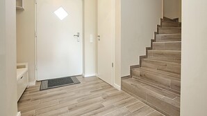 Un pasillo con una escalera de madera, una puerta blanca con un ventanal y un estante pequeño.