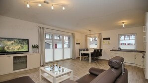Una sala de estar moderna con un sofá, una mesa de centro y un televisor. Hay un comedor con una mesa y sillas, y una cocina con armarios y un ventanal.