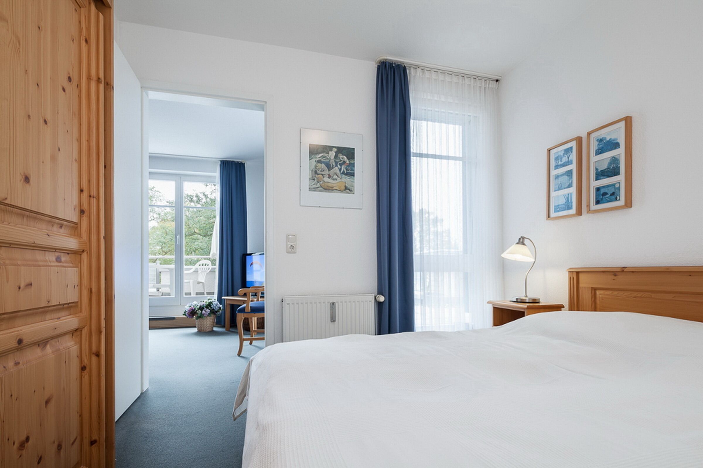 Habitación de hotel con una cama grande, un armario de madera, un escritorio con silla, una ventana con cortinas y cuadros en la pared.