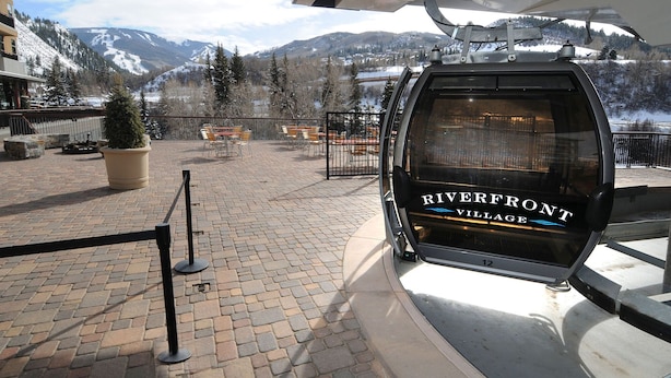 A ski lift with 'Riverfront Village' signage, a paved area with benches, and a view of snow-capped mountains. - Spacious Avon Retreat | Pool, Tennis, 2 Masters & Free Shuttle (Avon)