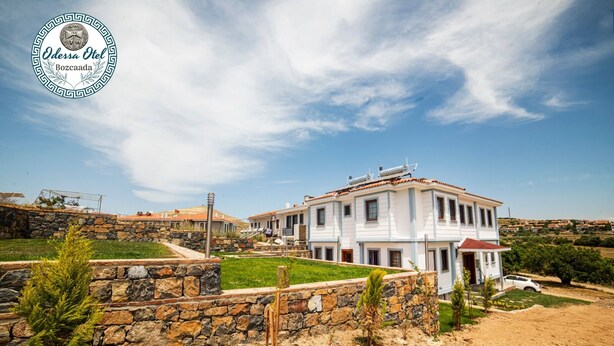 A large, two-story building with a white facade and red-tiled roof, surrounded by a stone wall and greenery. - Bozcaada Odessa  (Bozcaada)