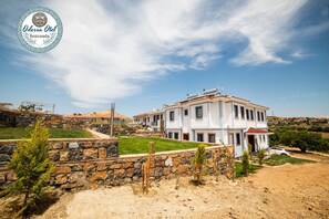 A large, two-story building with a white facade and red-tiled roof, surrounded by a stone wall and greenery. - Bozcaada Odessa  (Bozcaada)