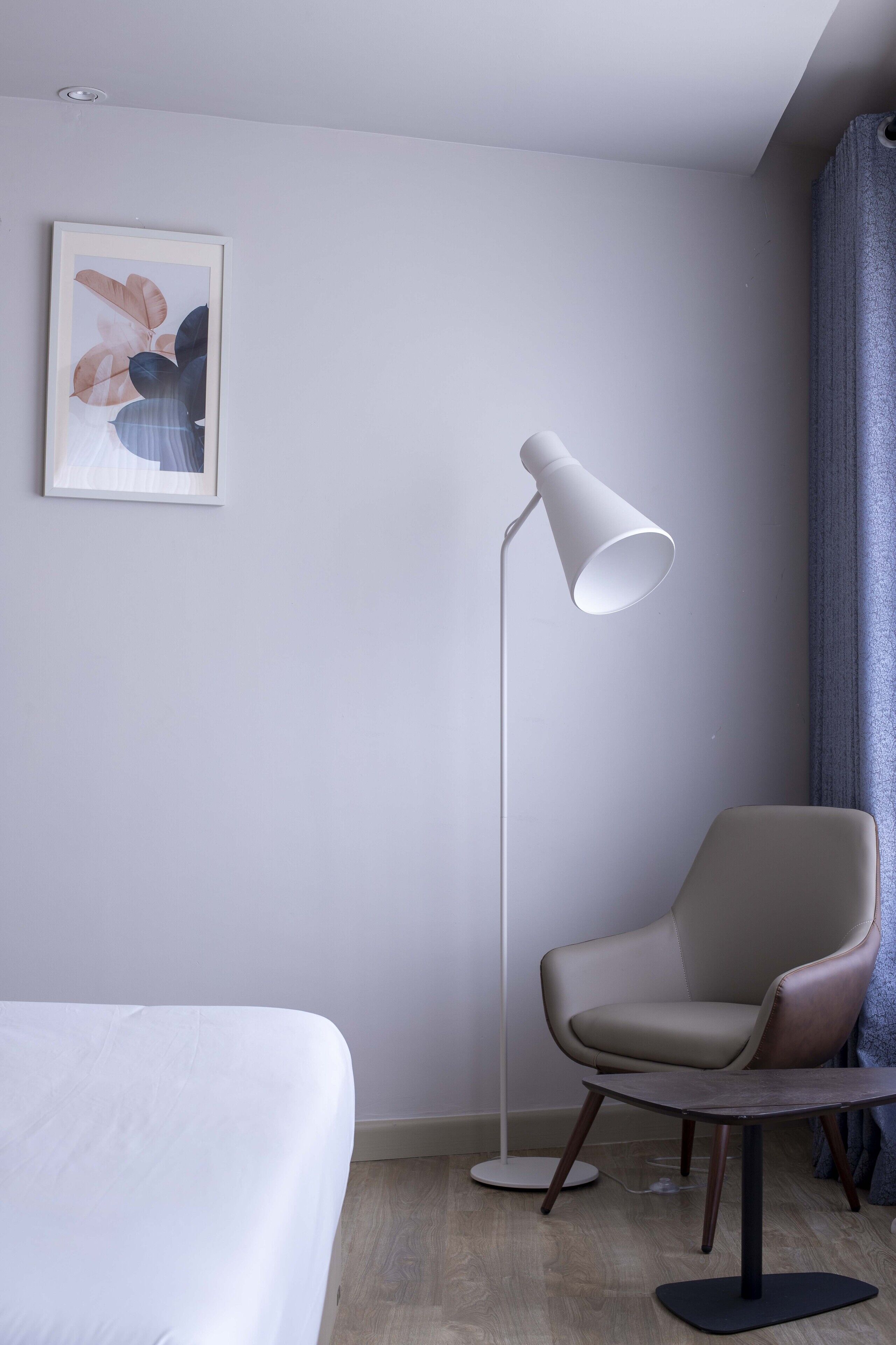 Een moderne hotelkamer met een bed, een stoel, een vloerlamp en een kunstwerk in een lijst aan de muur.