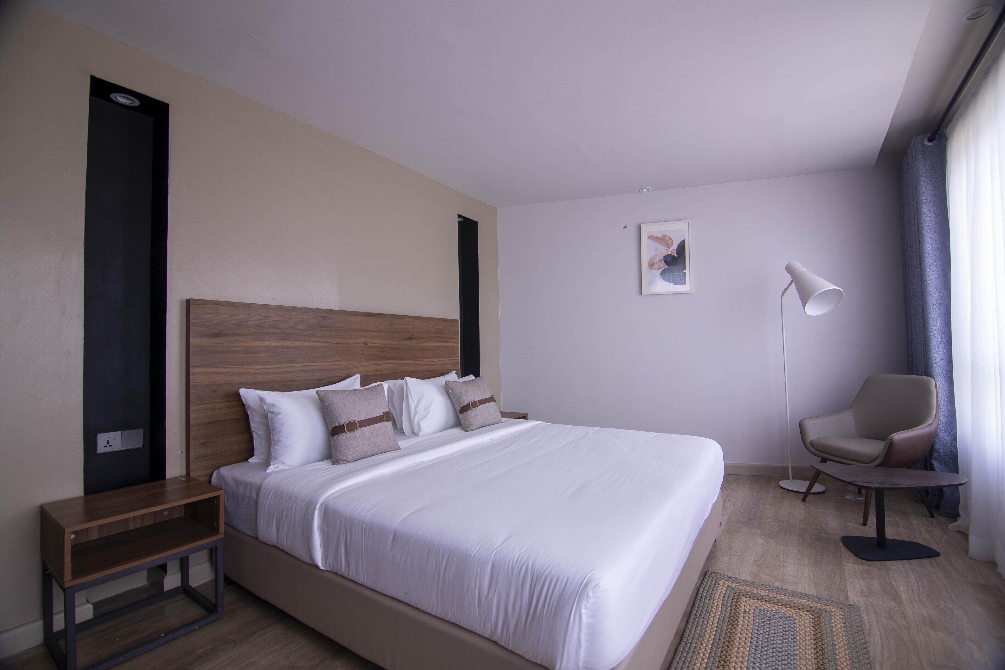 Une chambre d’hôtel avec un grand lit, une table de chevet, une chaise, une lampe sur pied et une petite œuvre d’art accrochée au mur.