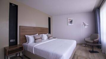 Une chambre d’hôtel avec un grand lit, une table de chevet, une chaise, une lampe sur pied et une petite œuvre d’art accrochée au mur.
