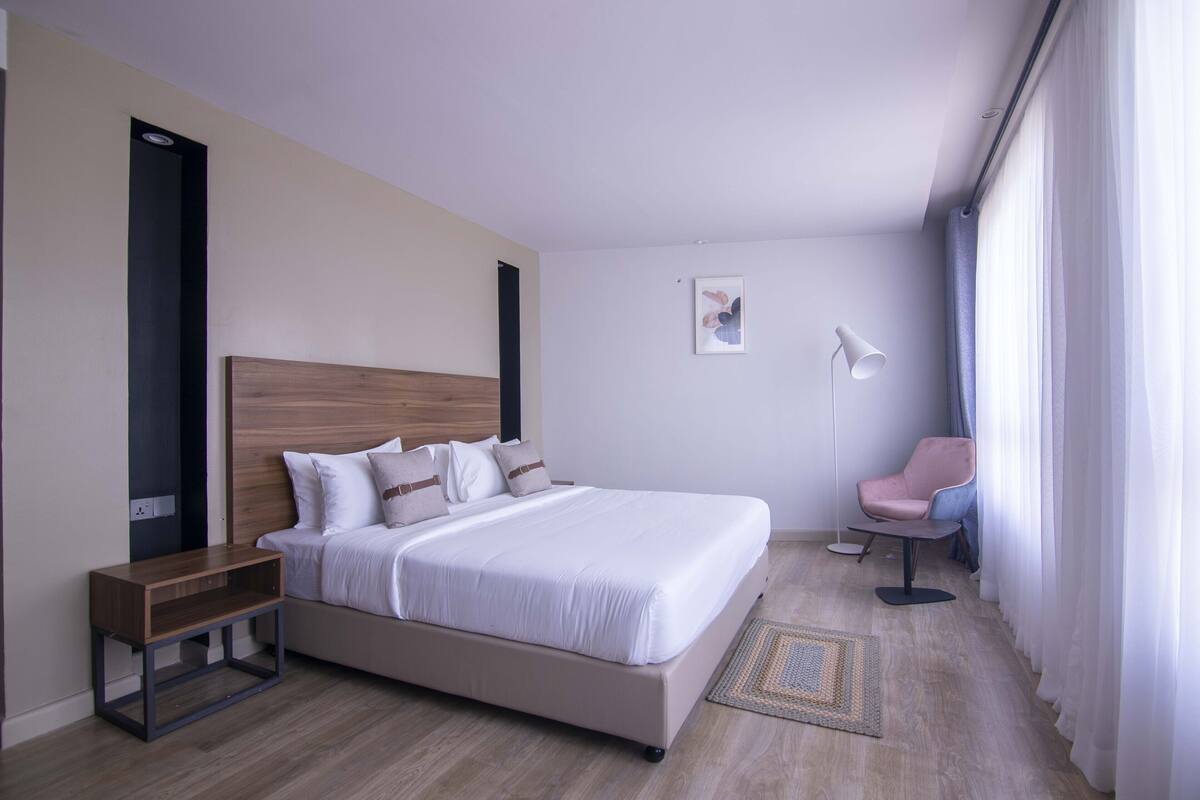 A modern hotel room with a large bed, a bedside table, a chair, and a floor lamp.