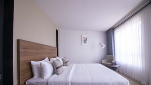A modern hotel room with a large bed, a wooden headboard, a bedside table, a chair, and a window with curtains.