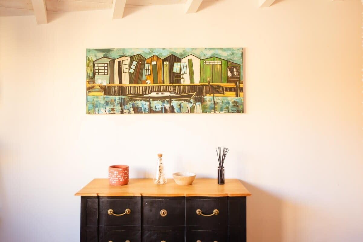 A painting of colorful houses by a water body on a wall above a wooden cabinet with drawers.