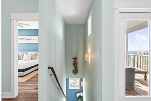 A hallway with a staircase leading to a bedroom with a bed and a window, a decorative wall hanging, and a view of a balcony with outdoor furniture. - A Modern Coastal Retreat Steps from the Gulf (Cape San Blas)