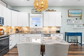 A modern kitchen with white cabinets, a granite countertop, a central island with chairs, and a hanging light fixture. - A Modern Coastal Retreat Steps from the Gulf (Cape San Blas)