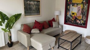 Ein Wohnzimmer mit einem beigen Sofa, roten Kissen, einem kleinen Couchtisch aus Holz, einer Topfpflanze und einem gerahmten Kunstwerk an der Wand.