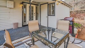 Een terras met een glazen tafel, rieten stoelen, een kleine schuur en een barbecue.