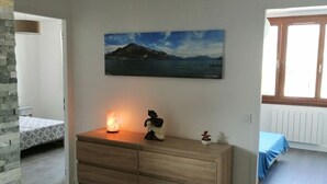 Ein Zimmer mit einem hölzernen Schminktisch, einer brennenden Kerze, einem Gemälde von einer Berglandschaft und einem Bett mit blauer Bettdecke.