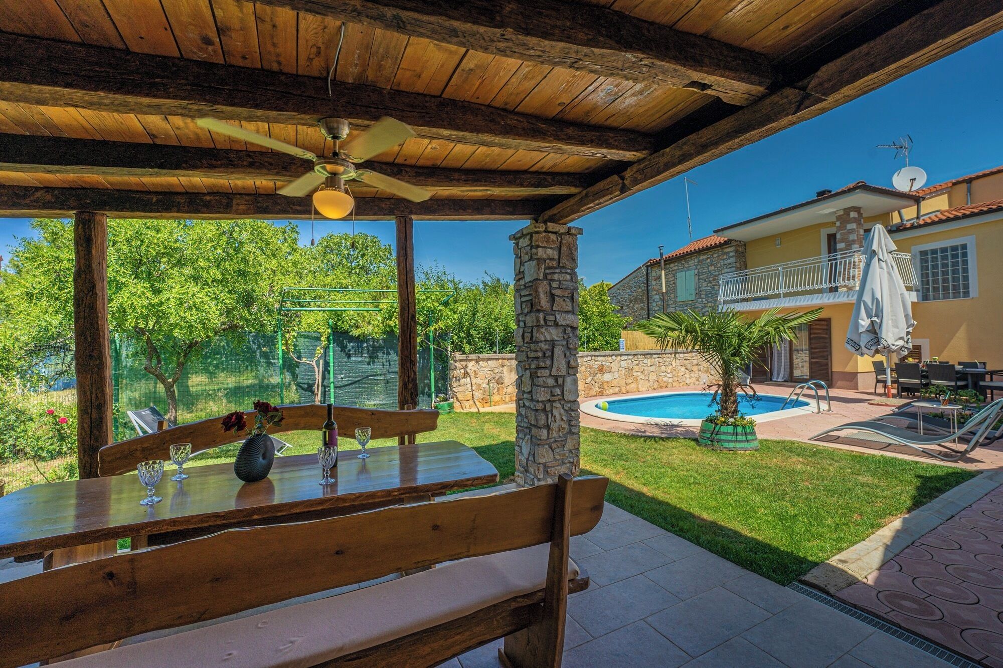 A wooden outdoor seating area with a bench and table, a stone pillar, a pool, and a building with a balcony and outdoor furniture.