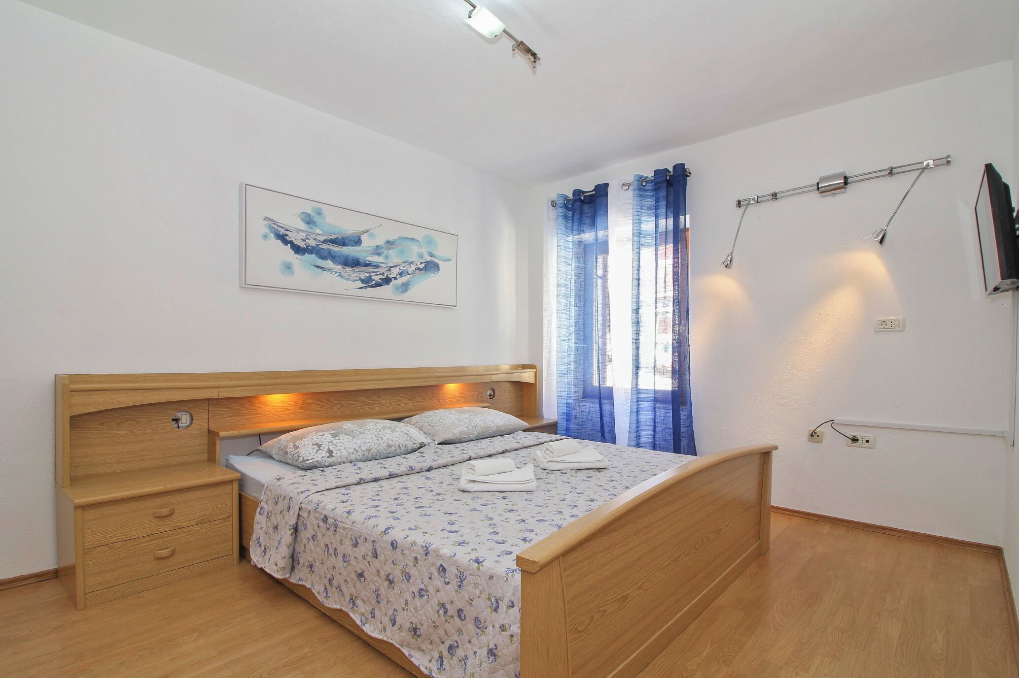 Un dormitorio con una cama de madera, una mesita de noche, un televisor montado en la pared y un cuadro.
