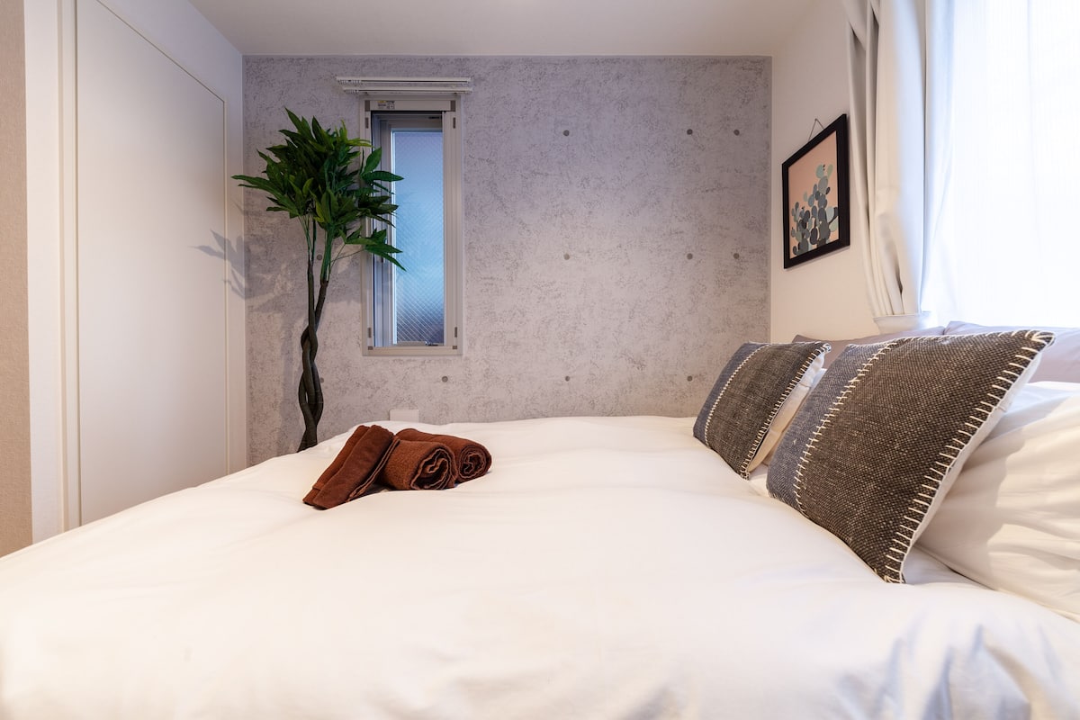 A neatly made bed with white linens and brown pillows, a potted plant, and a window with blinds.