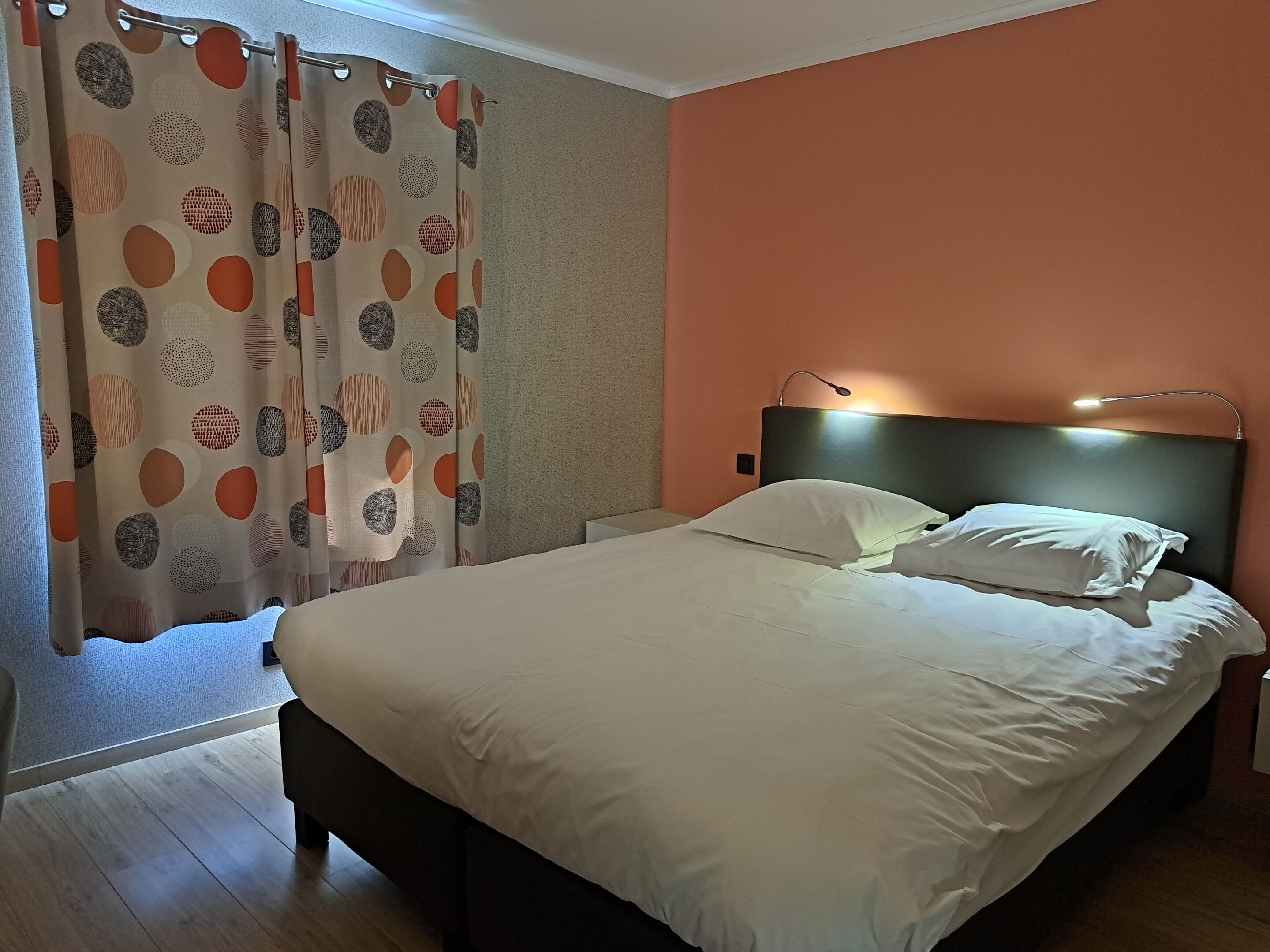 Een keurig opgemaakt bed met witte lakens, een hoofdbord met een lamp en een patroon gordijn voor een raam.