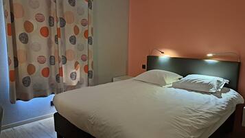 Een keurig opgemaakt bed met witte lakens, een hoofdbord met een lamp en een patroon gordijn voor een raam.