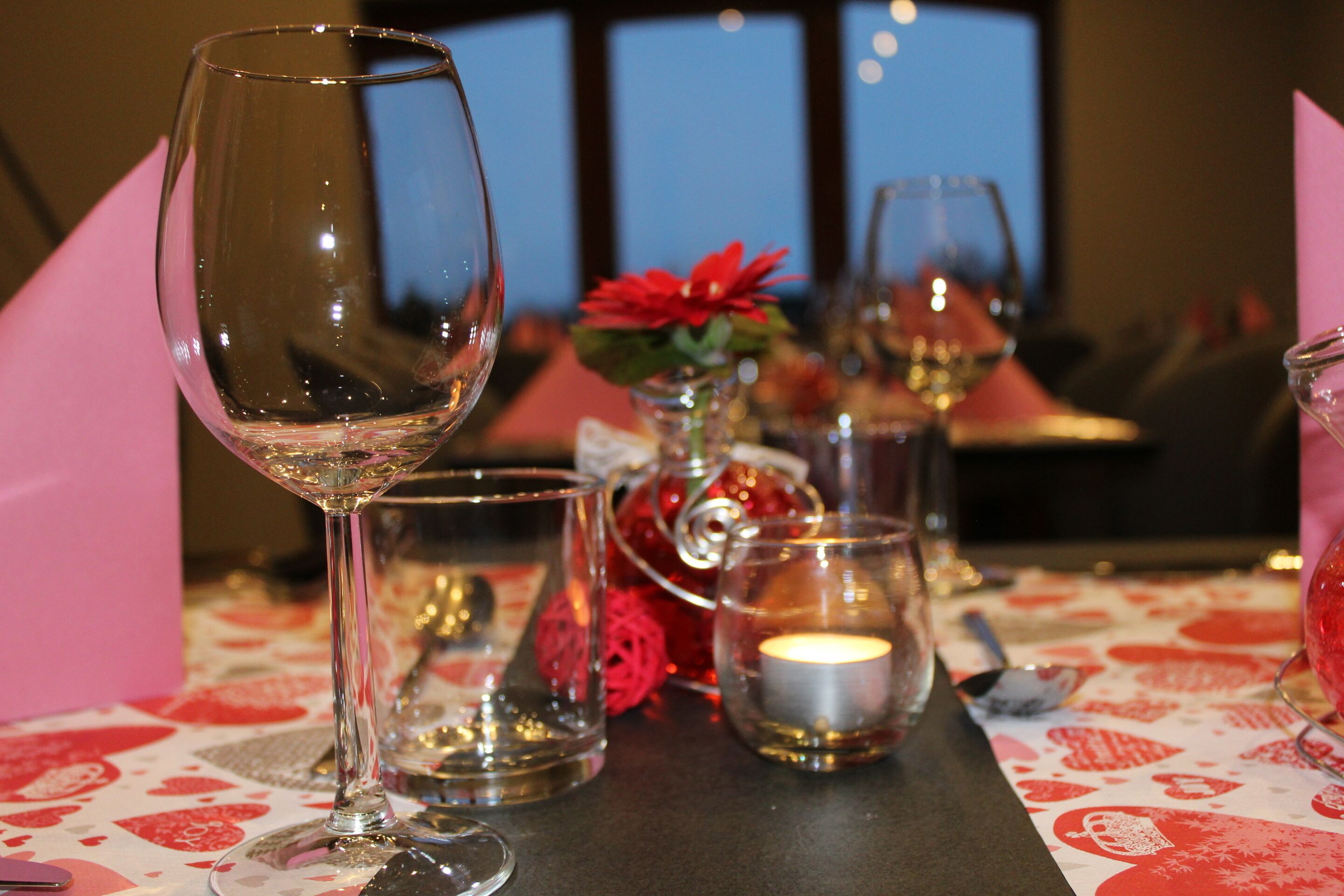 Une table dressée avec des verres à vin, une bougie et un vase décoratif contenant une fleur rouge.
