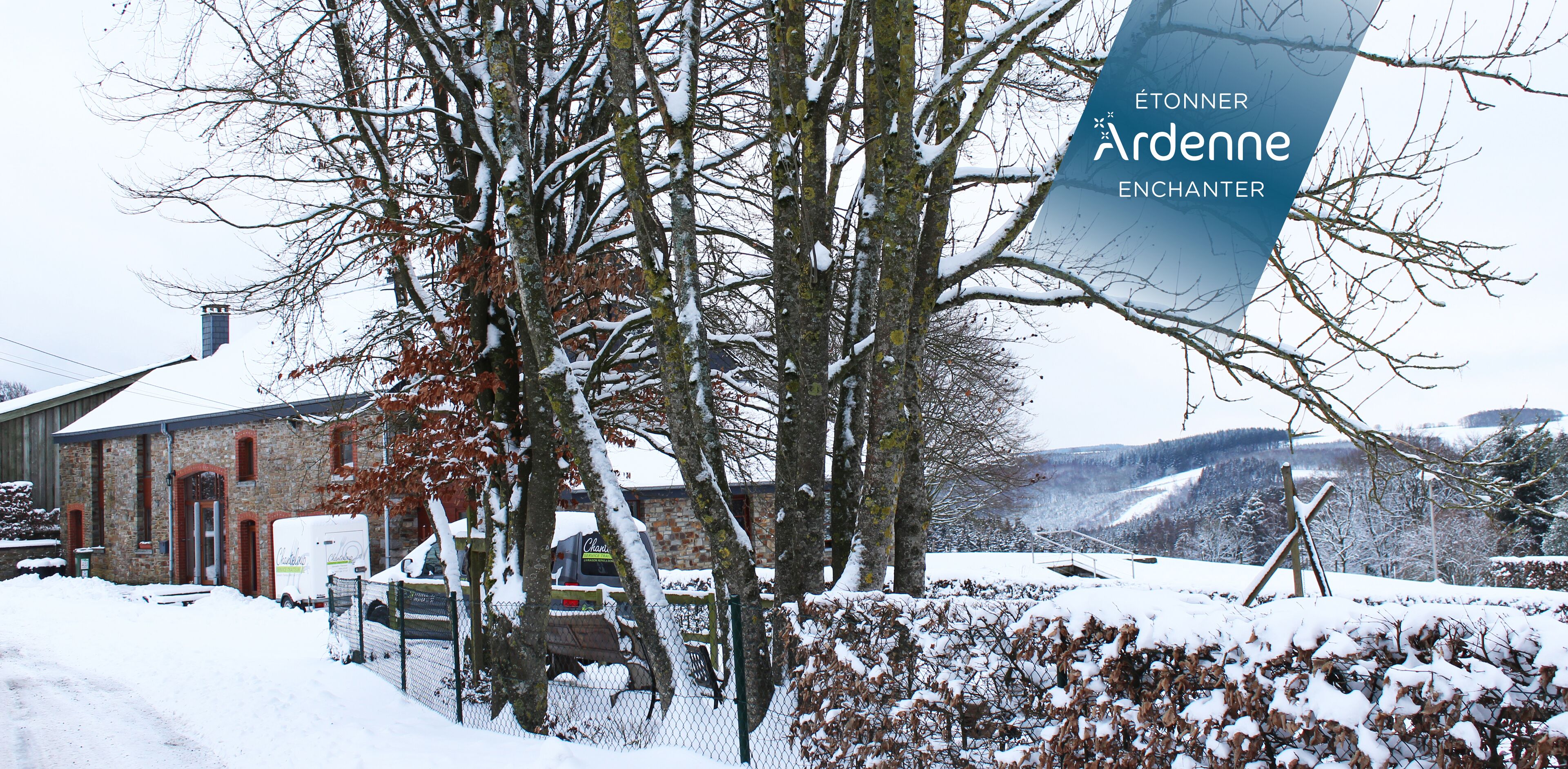 Un paysage enneigé avec un bâtiment en briques, un fourgon garé et une enseigne sur laquelle on peut lire : « ÉTONNER ARDENNE ENCHANTER ».