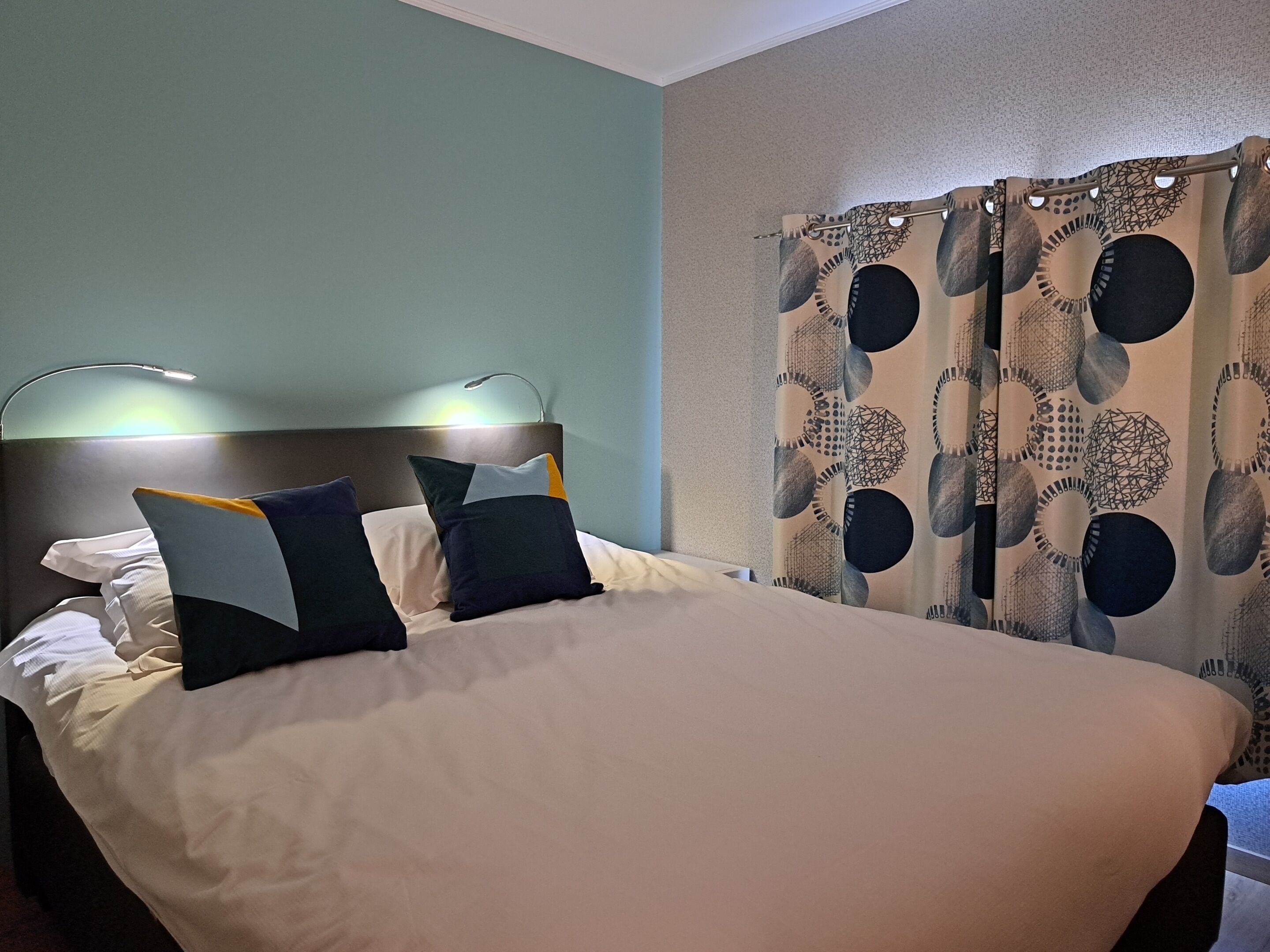 Un lit bien fait, avec du linge de lit blanc et deux oreillers, une tête de lit dotée d’un luminaire et un rideau orné d’un motif géométrique.