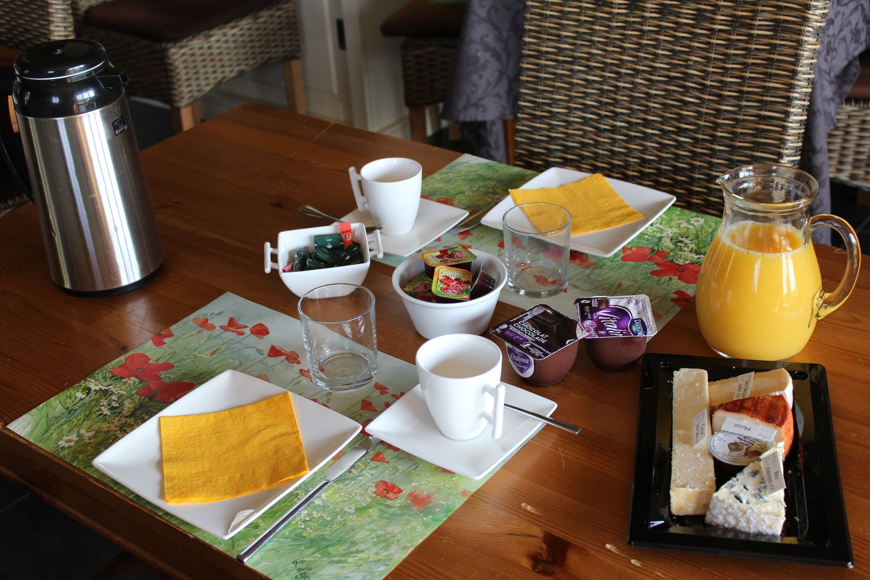 Une table de petit-déjeuner avec un thermos, des tasses, un pichet de jus d’orange et une assiette de fromage et de crackers.