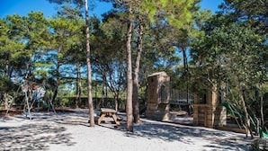 Un espace extérieur sablonneux avec une balançoire en bois, une structure en pierre et de grands arbres.