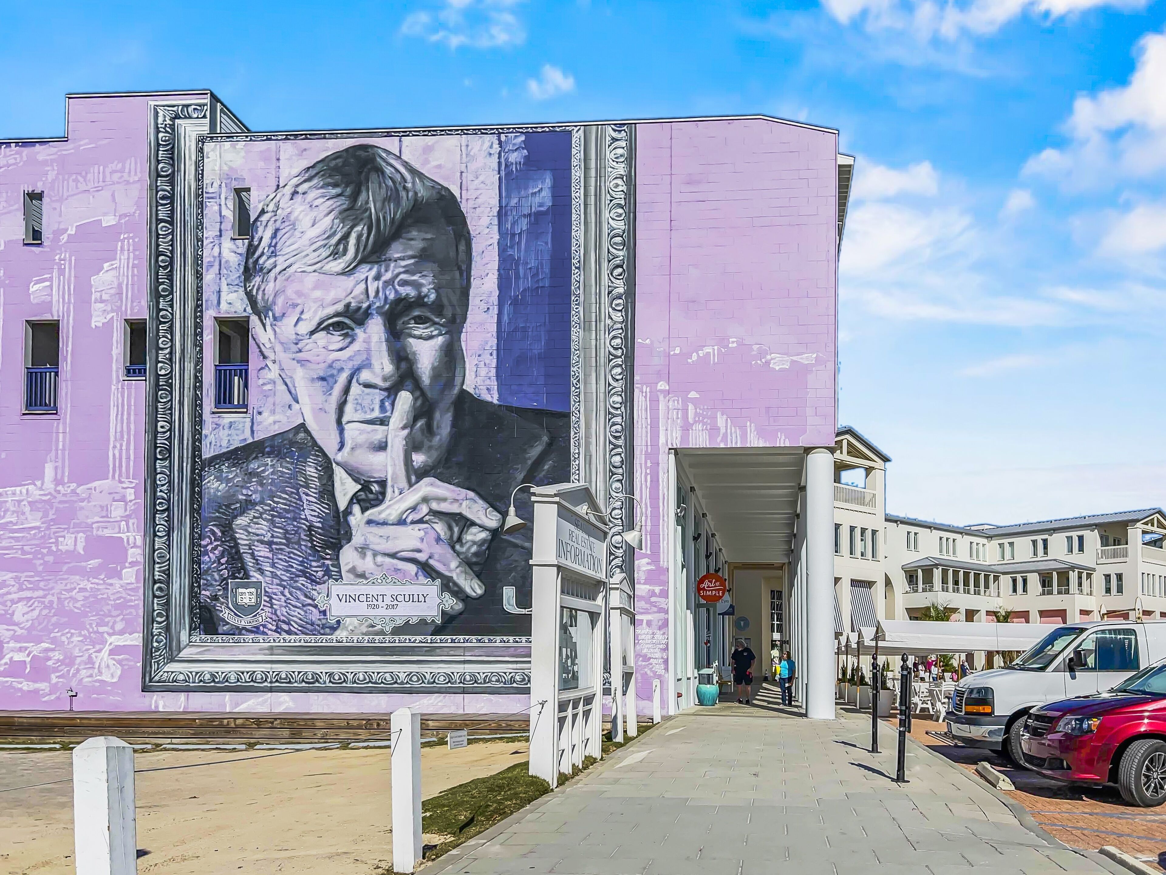 A building with a mural of a man in a suit and tie, a sign reading "Vincent Scully 1920-2017," and a "REAL ESTATE INFORMATION" sign.