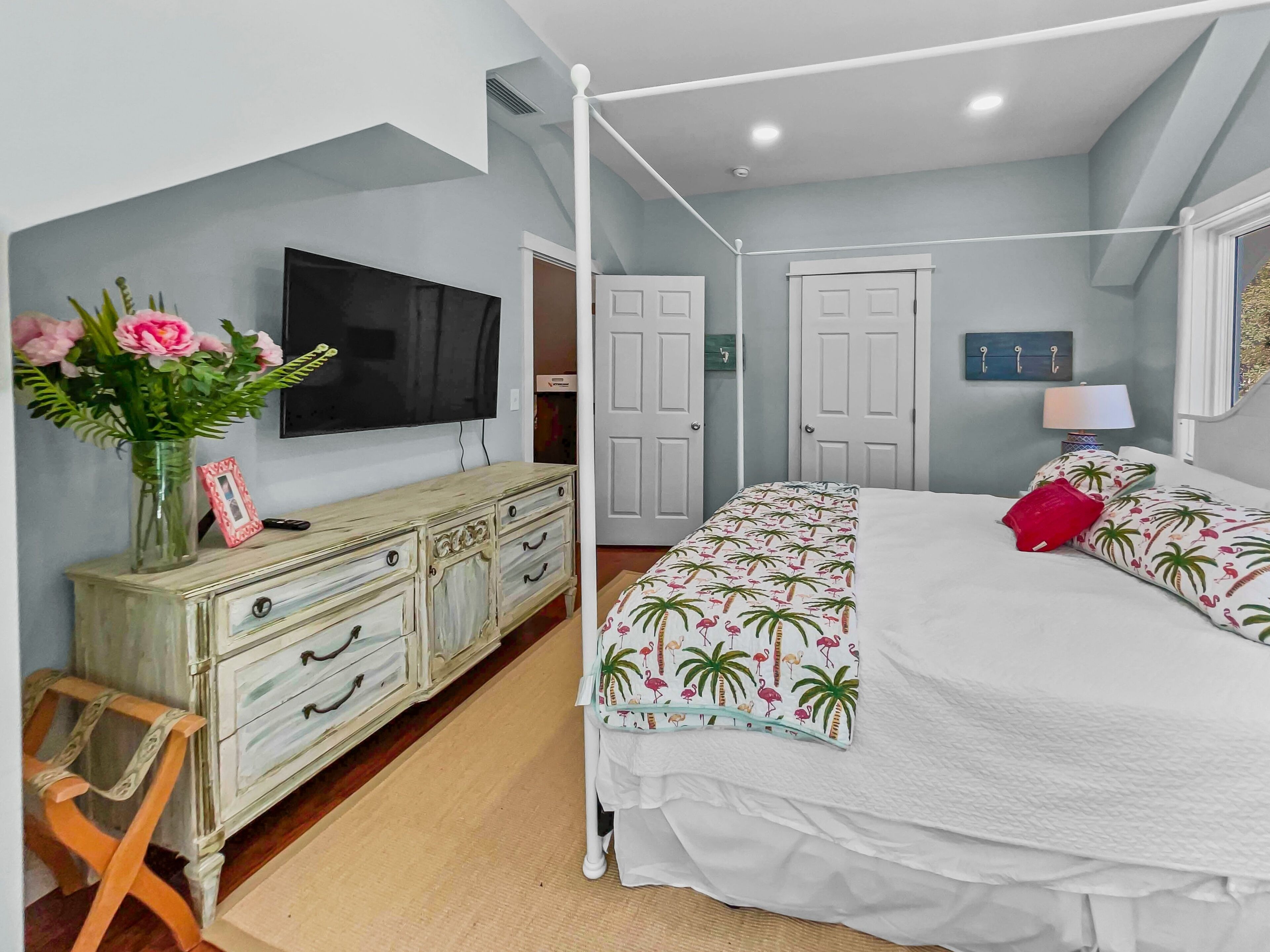 A bedroom with a four-poster bed, a television mounted on the wall, a dresser with drawers, and a vase with flowers.
