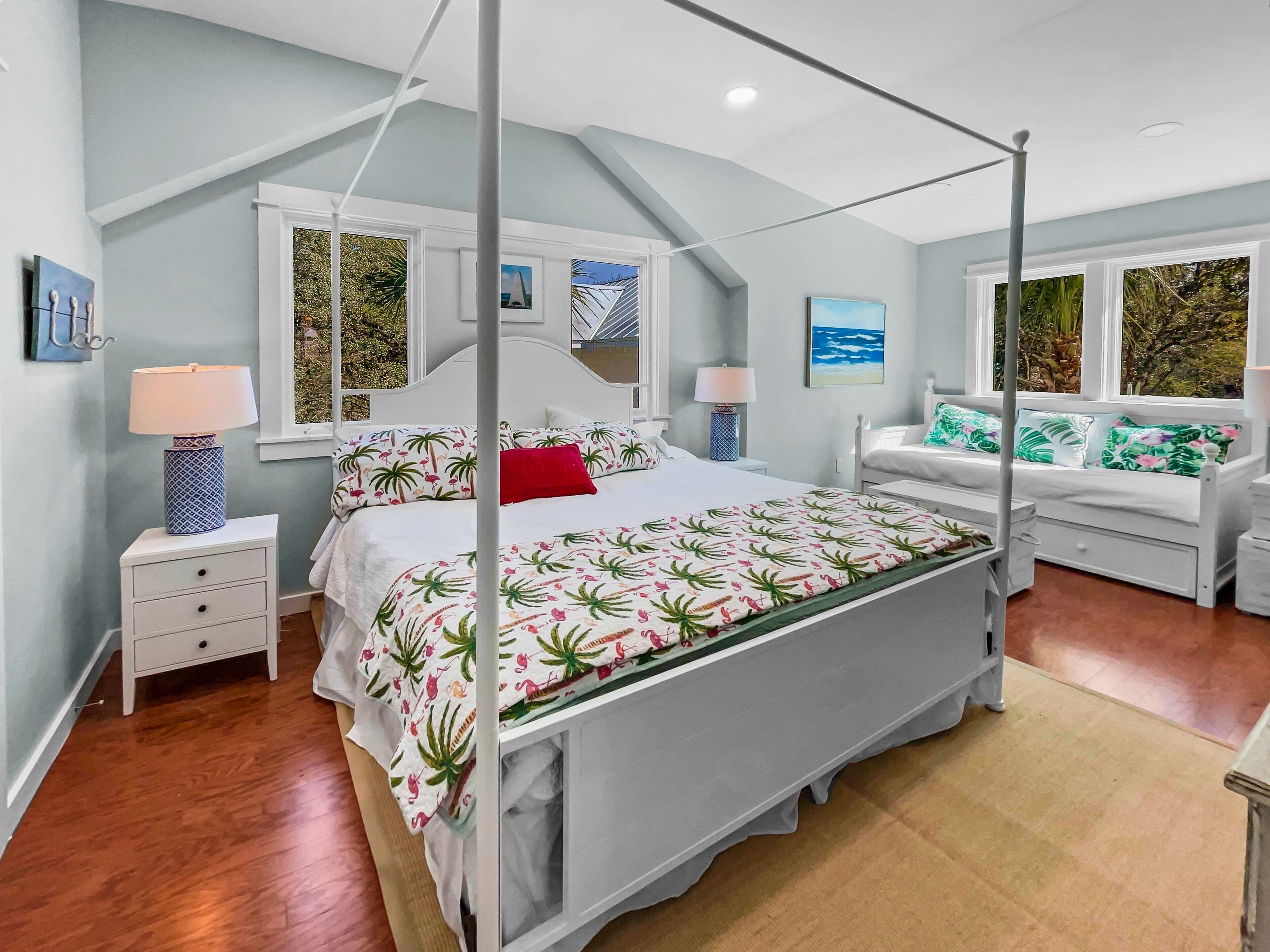A four-poster bed with a tropical-themed bedspread, white nightstands with lamps, and a white sofa with green and pink pillows.