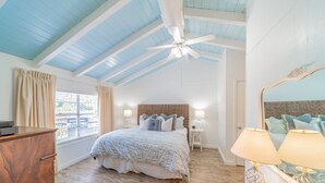 Une chambre avec un grand lit, un ventilateur de plafond, une commode en bois, une fenêtre avec des rideaux et un miroir doté d’un cadre décoratif.