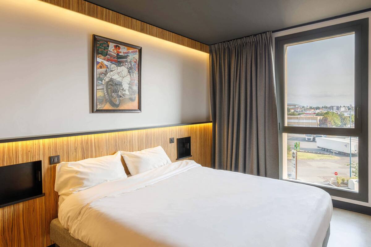 A hotel room with a large bed, a mounted TV, a window with curtains, and a city view.