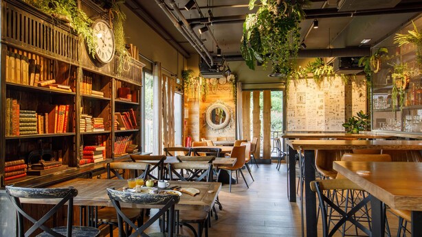 A cozy cafe with wooden tables and chairs, bookshelves filled with books, and hanging plants.